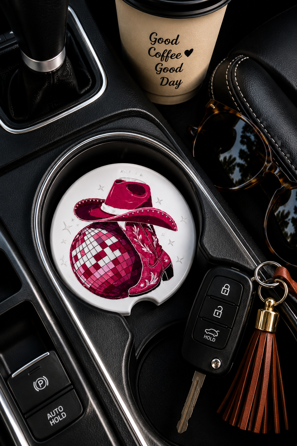 Disco Cowgirl Car Coaster sitting in a car cup holder next to a coffee cup, sunglasses, and keys, showing the pink disco ball, cowboy hat, and boots design.
