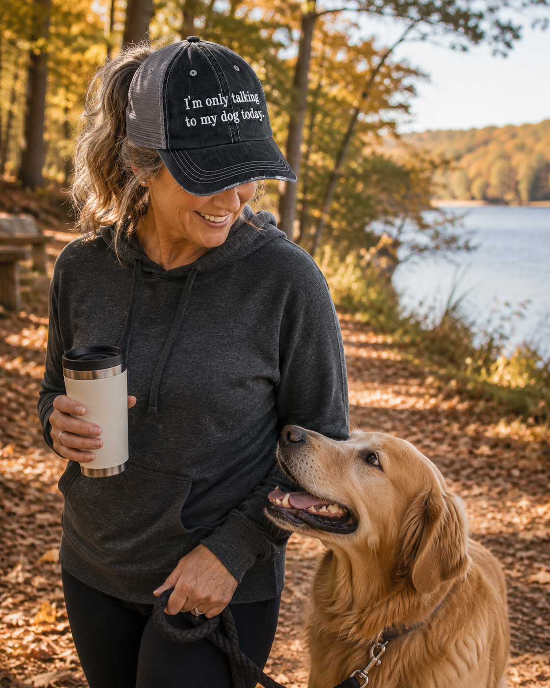 A woman wearing a black trucker hat with the text 'I'm only talking to my dog today.' stands outdoors by a lake with a golden retriever dog beside her, holding a travel mug.