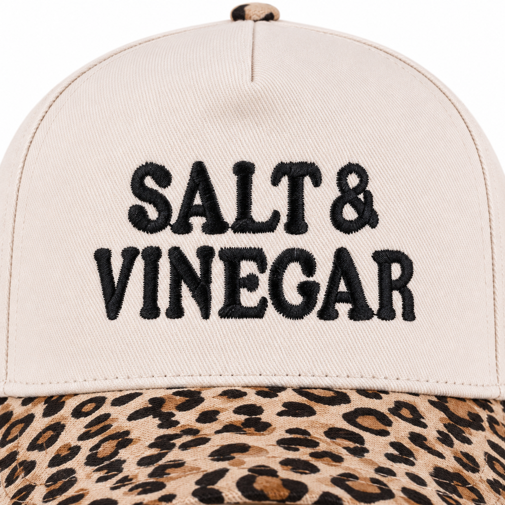 Close-up of Salt & Vinegar Two-Tone Baseball Cap in Leopard showing bold black raised embroidered Salt & Vinegar block lettering on cream structured cotton crown with tan and brown leopard print bill visible along bottom edge on white background
