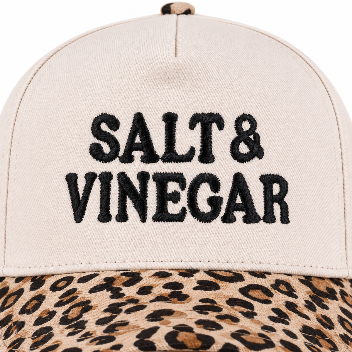 Close-up of Salt & Vinegar Two-Tone Baseball Cap in Leopard showing bold black raised embroidered Salt & Vinegar block lettering on cream structured cotton crown with tan and brown leopard print bill visible along bottom edge on white background
