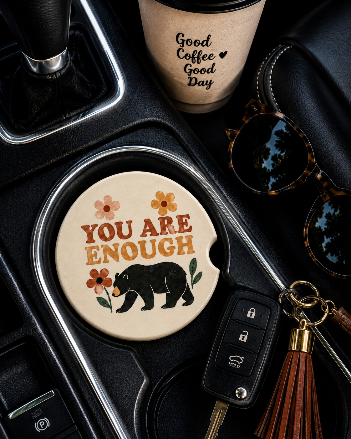 You Are Enough ceramic car coaster in cup holder — black bear and wildflower inspirational design, shown in car console with coffee cup and sunglasses
