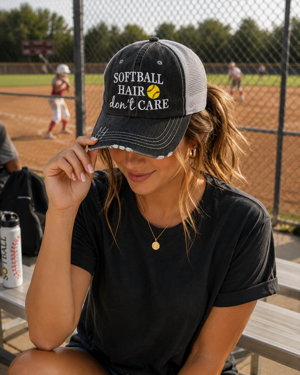 A woman sitting at a baseball field bench wearing a black and gray trucker hat with the text 'SOFTBALL HAIR don't CARE' and a yellow softball graphic. The background shows a chain-link fence and a softball field with players.