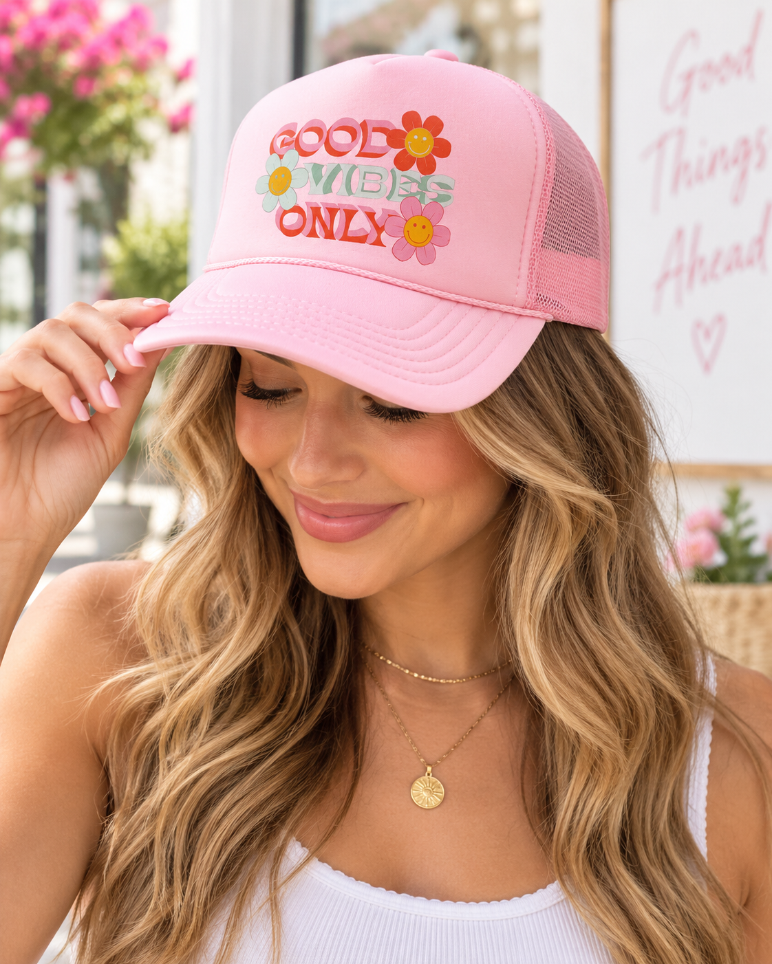 Woman wearing Good Vibes Only Trucker Hat with pink front and pink mesh back showing retro Good Vibes Only lettering with smiley face daisy flowers with white tank and outdoor floral backdrop
