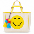 Smiley Face Tote Bag front view with natural canvas body, large textured punch needle yellow smiley face, yellow striped handles, yellow trim, and colorful pink yellow and blue pom-pom tassel accent on white background
