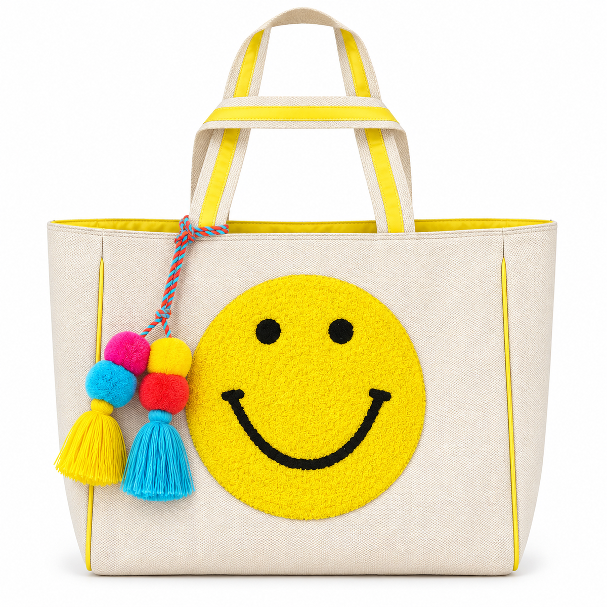 Smiley Face Tote Bag front view with natural canvas body, large textured punch needle yellow smiley face, yellow striped handles, yellow trim, and colorful pink yellow and blue pom-pom tassel accent on white background
