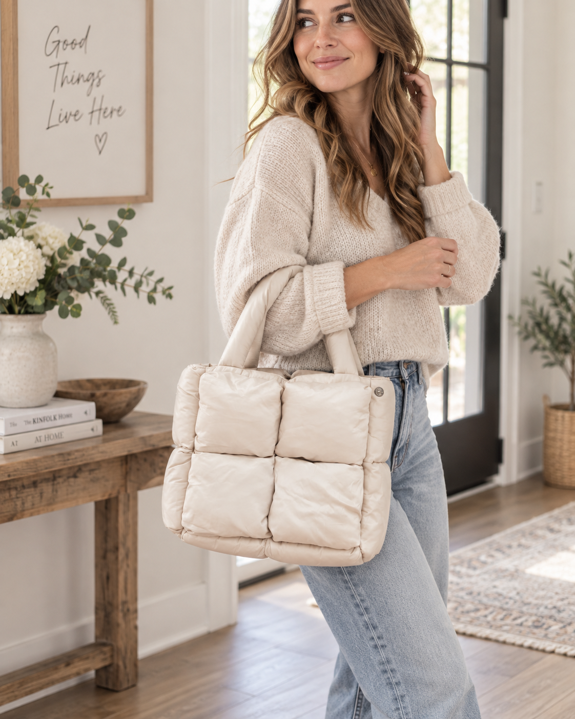 Woman carrying Sand Mini Quilted Puffer Tote by top handles showing soft sand neutral puffer body with six padded square panels, padded handles, and adjustable shoulder strap with cream sweater and jeans in bright farmhouse entryway
