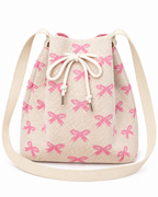 Coquette Bow Crossbody Bucket Bag front view showing cream woven knit fabric with allover pink bow pattern, cream rope drawstring closure with silver aglet tips, and wide flat cream crossbody strap with cream fabric attachment tabs on white background
