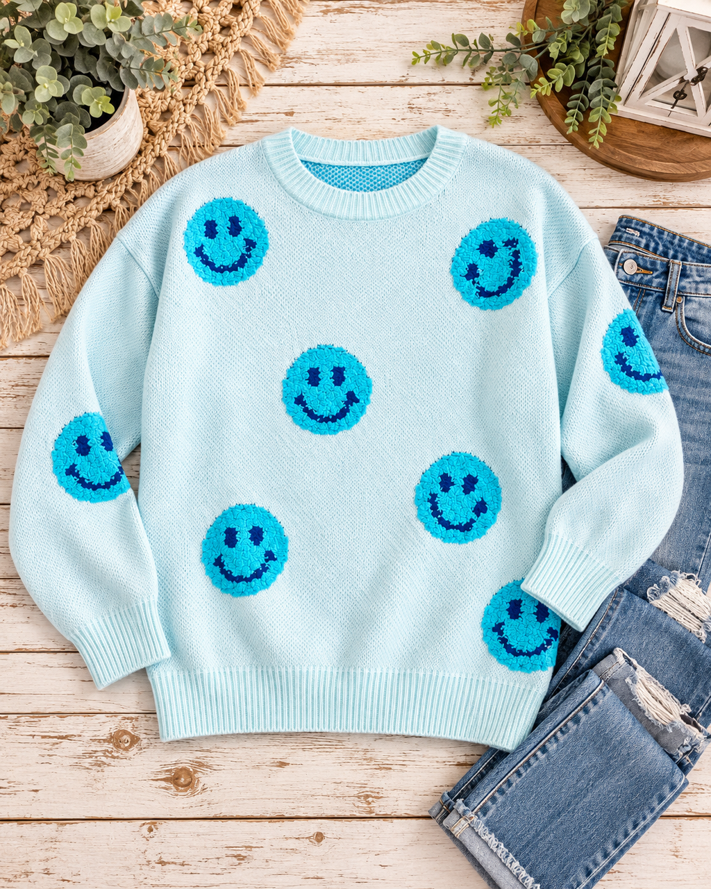 Light blue happy face knit sweater styled with distressed jeans, greenery, a fringed textile, and a lantern on rustic wood
