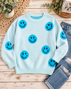 Light blue happy face knit sweater styled with distressed jeans, greenery, a fringed textile, and a lantern on rustic wood
