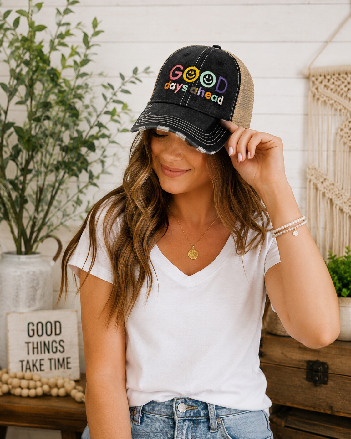 A woman wearing a distressed black and tan trucker hat with colorful 'GOOD days ahead' embroidery and smiley face accents, styled in a cozy home setting.
