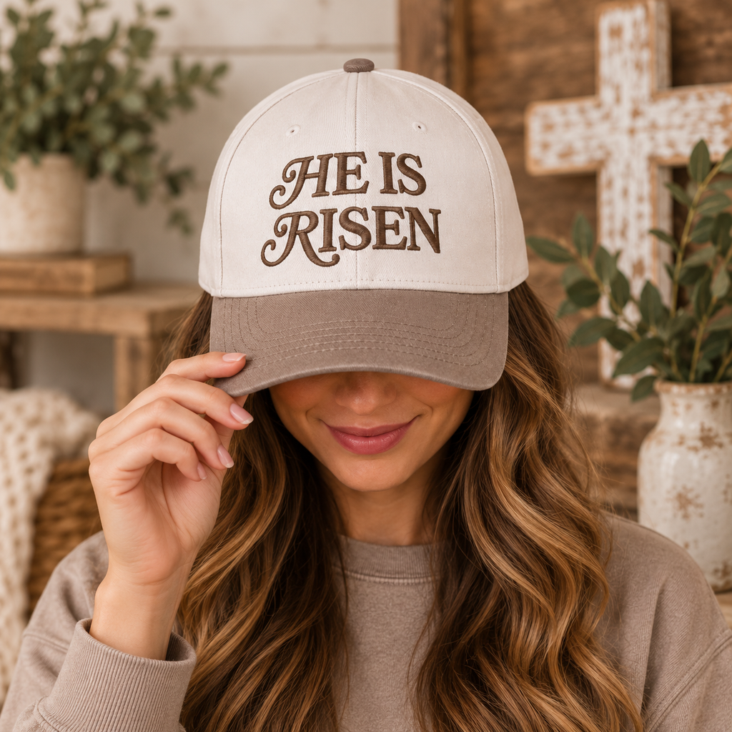 Brunette woman with long wavy highlighted hair in mocha crewneck smiling while pulling brim of He Is Risen Baseball Cap in Ash Mocha down over face, in farmhouse interior with wood cross and white vase with greenery in background
