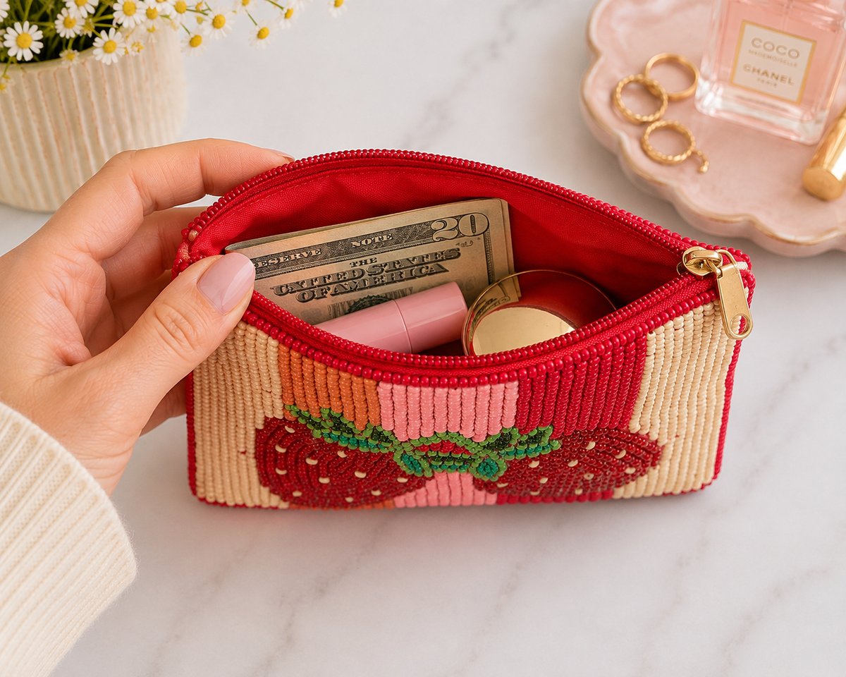 A woven cosmetic pouch with a strawberry design, held open by a hand. The pouch has a red interior, a red zipper, and a beige and pink exterior with a green embroidered strawberry motif.