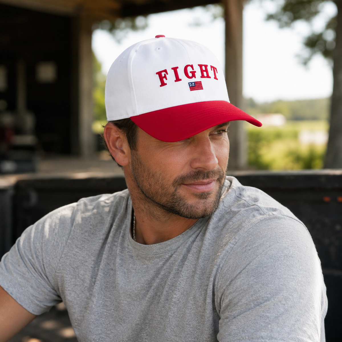 A man relaxes on an outdoor porch wearing a white and red Fight Baseball Cap with bold red embroidered "FIGHT" lettering and American flag accent, wearing a gray t-shirt with a rural backdrop.
