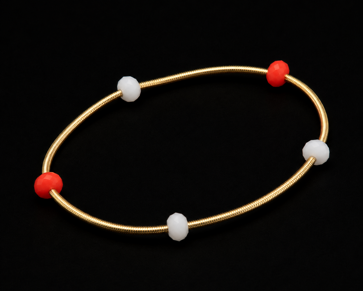 Gold coil stretch bracelet with red and white bead accents shown at an angle on black reflective surface
