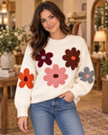Brunette woman smiling in cream knit sweater with five oversized 3D knit daisy floral appliqués in burgundy, brown, pink, orange, and gray, paired with blue jeans in a warm boutique interior with gold mirrors and florals
