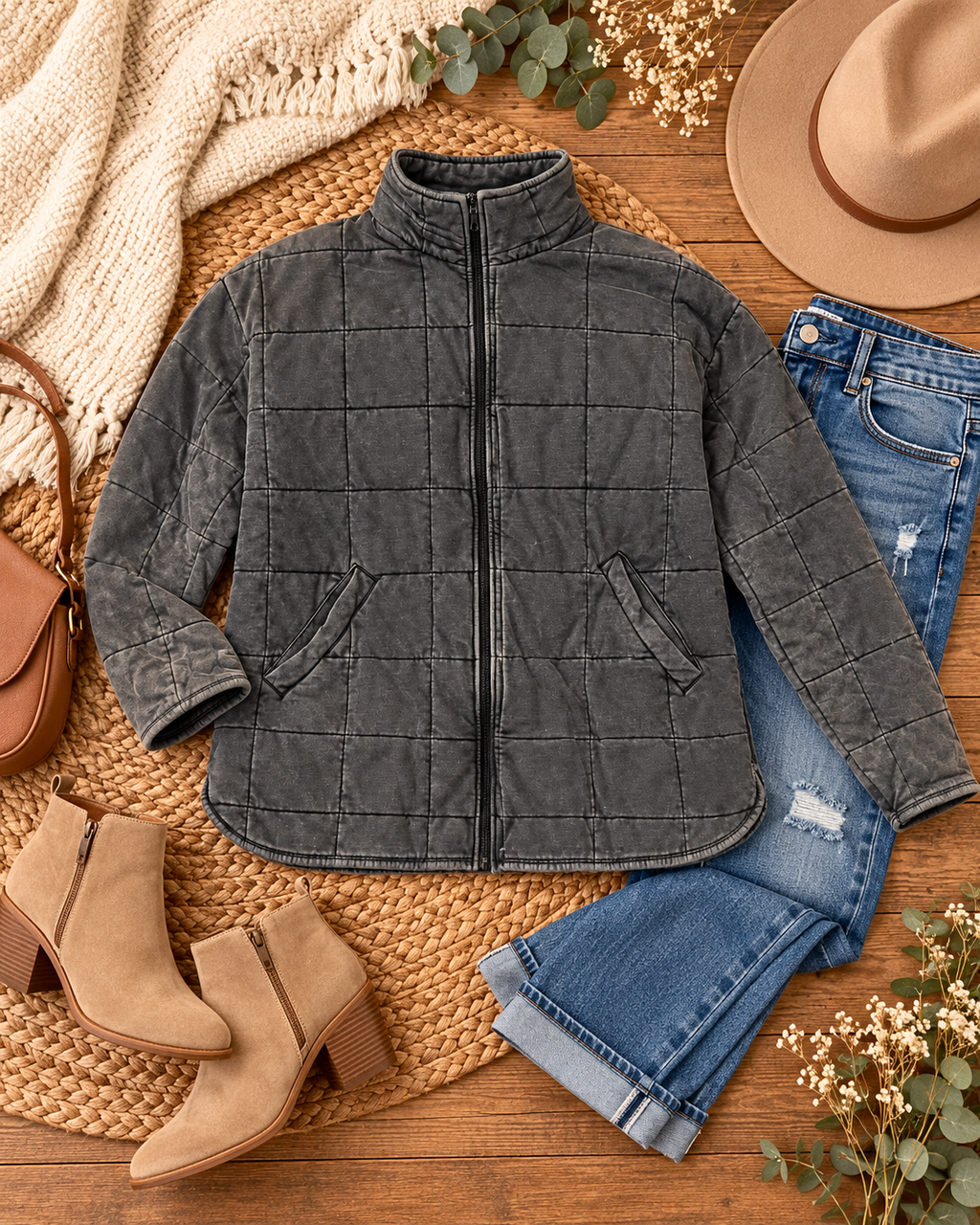 Charcoal Garment Washed Quilted Jacket styled in flat lay on woven rattan surface with distressed blue jeans, tan suede block-heel ankle booties, tan structured crossbody bag, beige felt fedora, cream knit blanket, and eucalyptus and baby's breath stems
