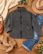 Charcoal Garment Washed Quilted Jacket styled in flat lay on woven rattan surface with distressed blue jeans, tan suede block-heel ankle booties, tan structured crossbody bag, beige felt fedora, cream knit blanket, and eucalyptus and baby's breath stems
