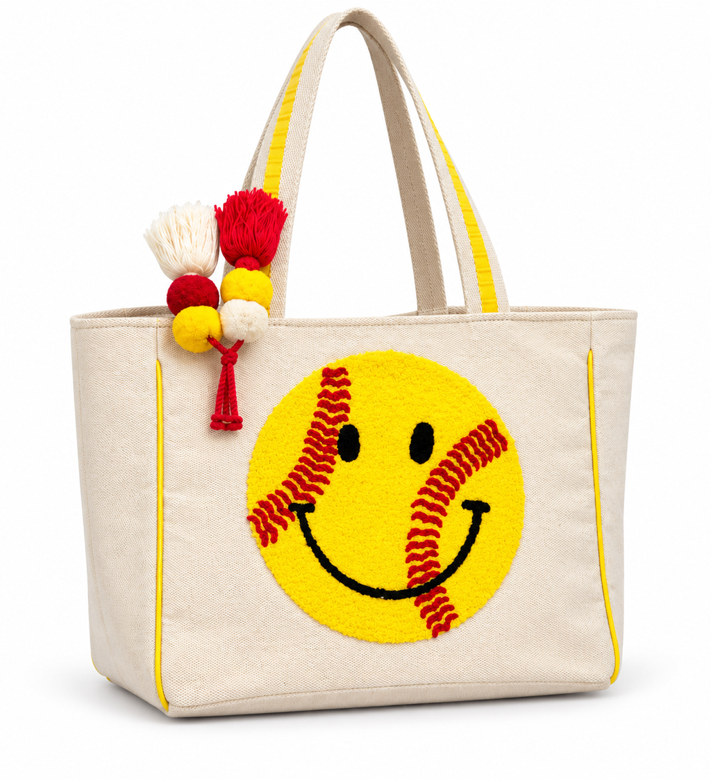 Softball Smiley Tote Bag three-quarter angle view showing natural canvas body, large yellow textured softball smiley face with red stitching, yellow trim handles, and red yellow and cream pom-pom tassel on white background
