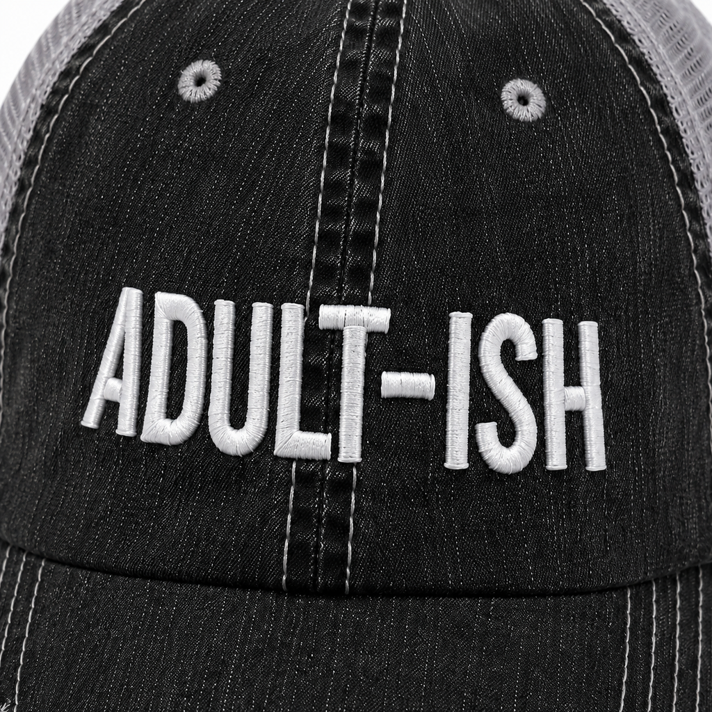 Close-up of a black distressed trucker hat with white embroidered text reading 'ADULT-ISH' on the front panel.