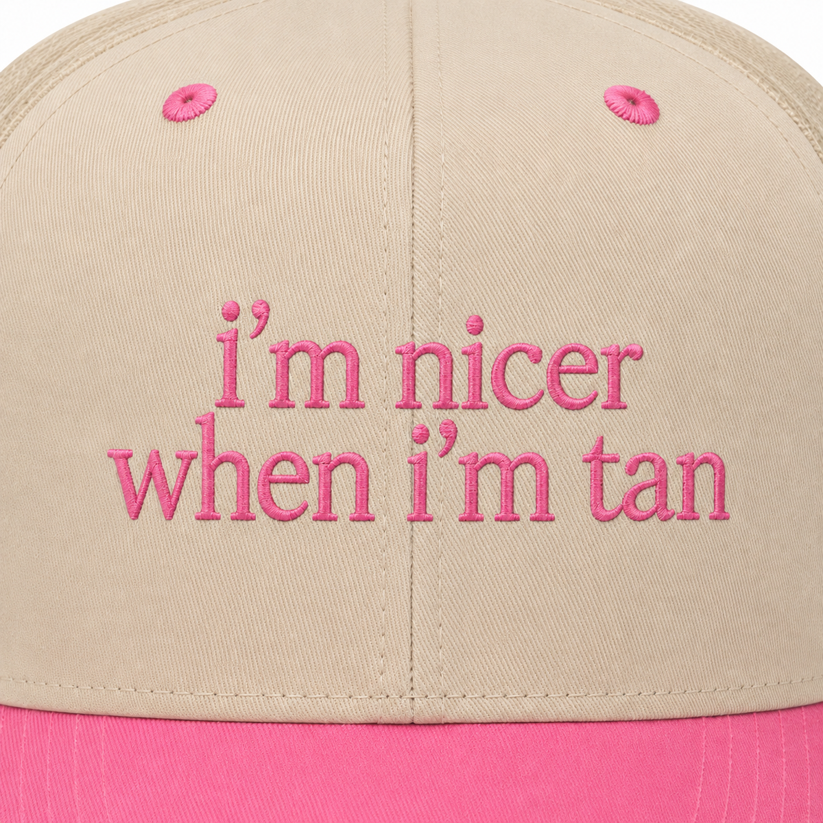 Close-up of soft pink embroidered "I'm Nicer When I'm Tan" lettering on a cream trucker hat front panel with pink brim.
