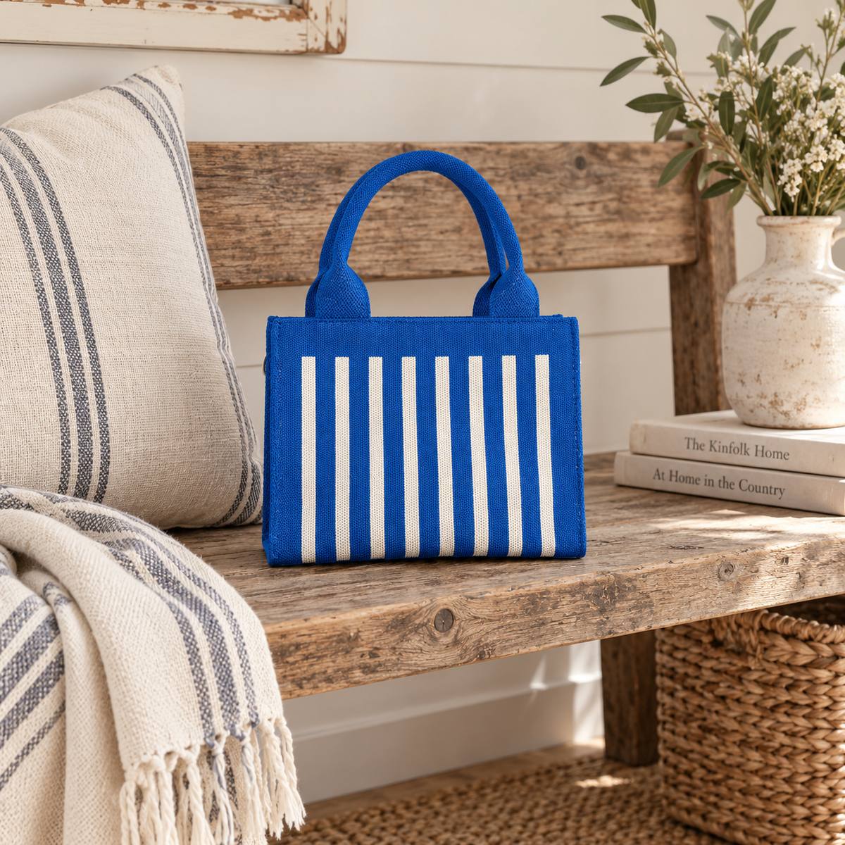 Mini Blue Striped Tote Handbag displayed on rustic wooden bench showing vivid cobalt blue structured woven body with cream vertical stripes and matching handles beside stacked books, ceramic vase with wildflowers, and striped throw in farmhouse setting

