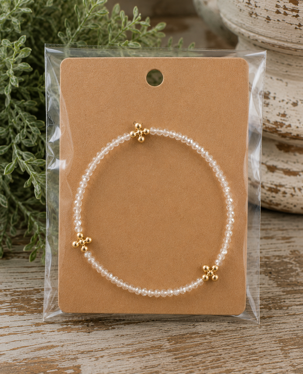 Clear beaded cross bracelet with gold cross accents on kraft card in clear packaging on rustic wood surface with chippy white pedestal and greenery
