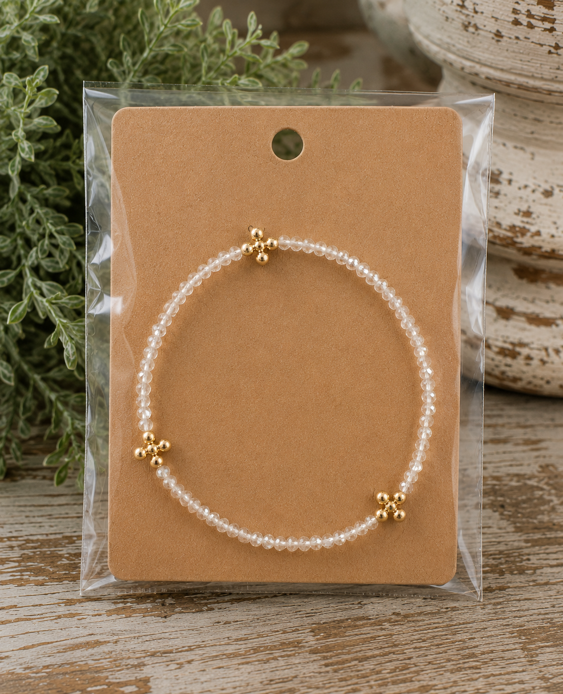 Clear beaded cross bracelet with gold cross accents on kraft card in clear packaging on rustic wood surface with chippy white pedestal and greenery
