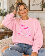 Seated model wearing a pink crewneck sweatshirt with white cowboy boots and hot pink detailing, paired with jeans
