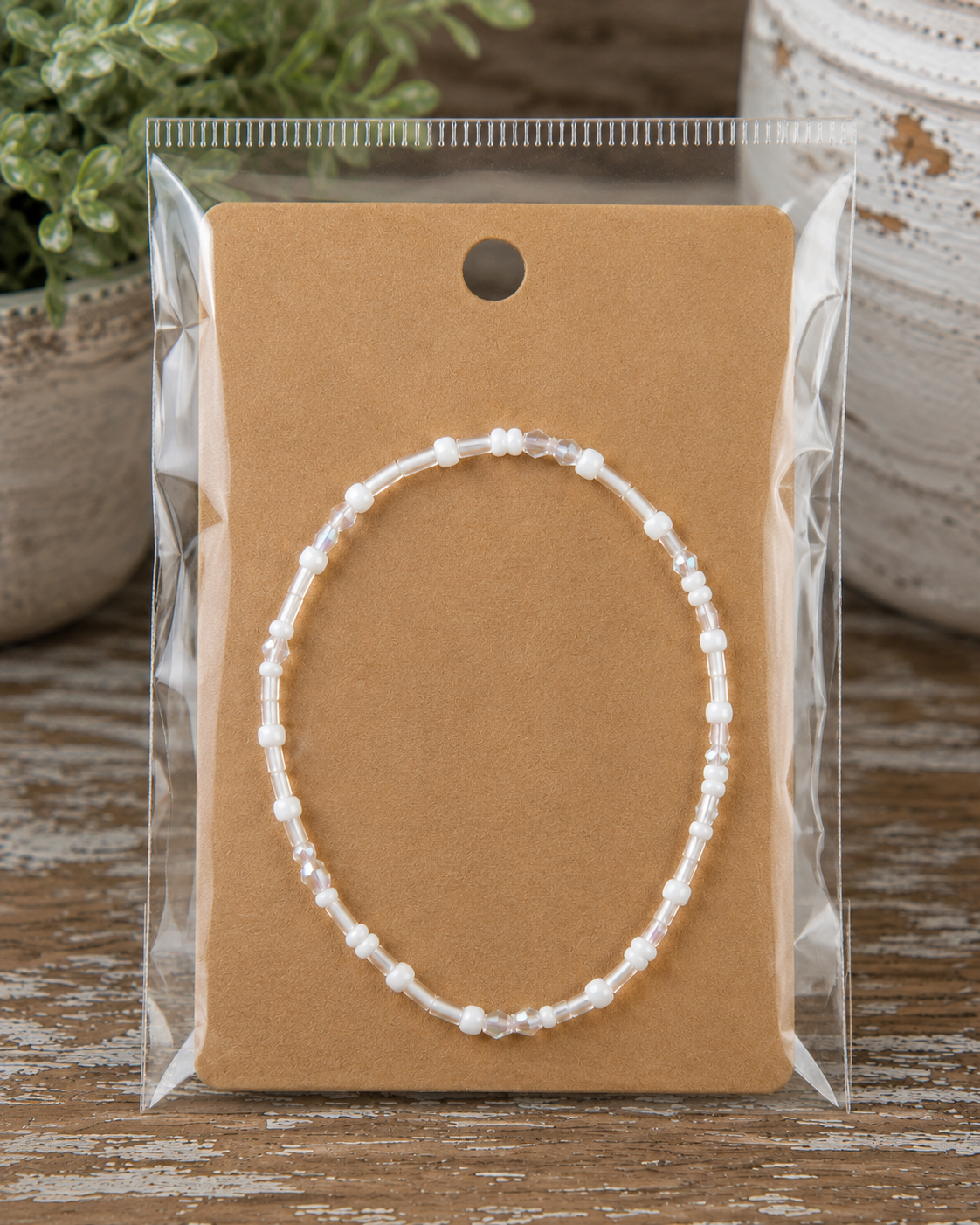 White and iridescent seed bead stretch bracelet on kraft card in clear packaging on rustic wood surface with chippy white pedestal and greenery backdrop
