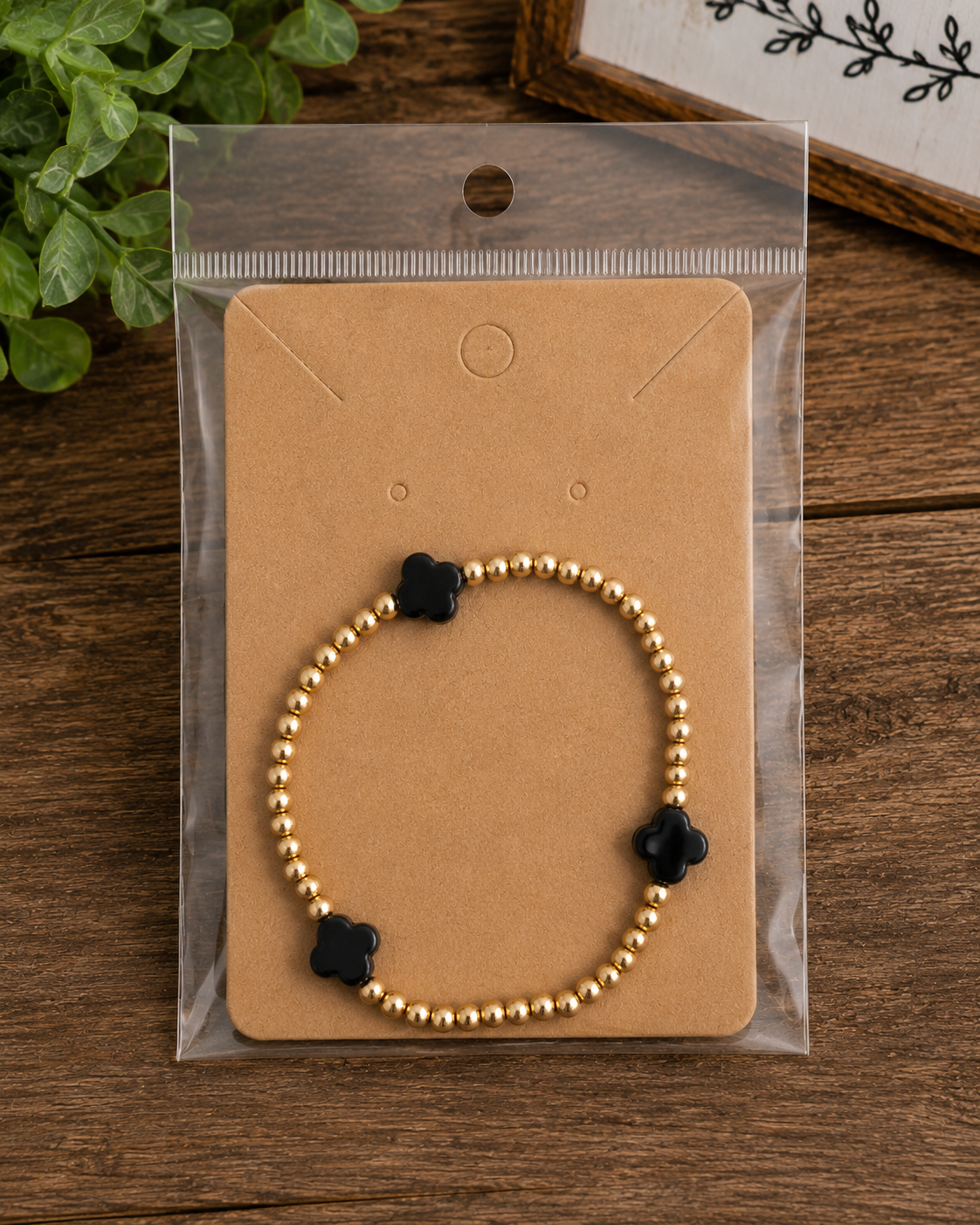 Gold beaded bracelet with black clover accents on kraft card in clear packaging on rustic wood surface with greenery and be still sign
