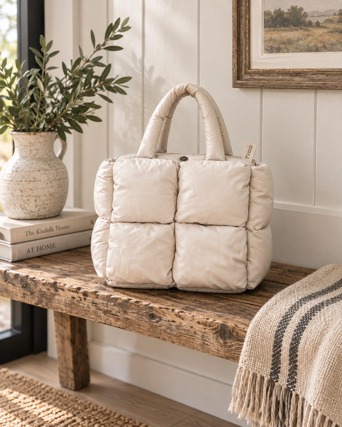 Sand Mini Quilted Puffer Tote displayed on rustic wooden bench showing soft sand neutral puffer body with six padded square panels and padded top handles beside ceramic vase with olive branches, stacked books, and woven throw in farmhouse setting

