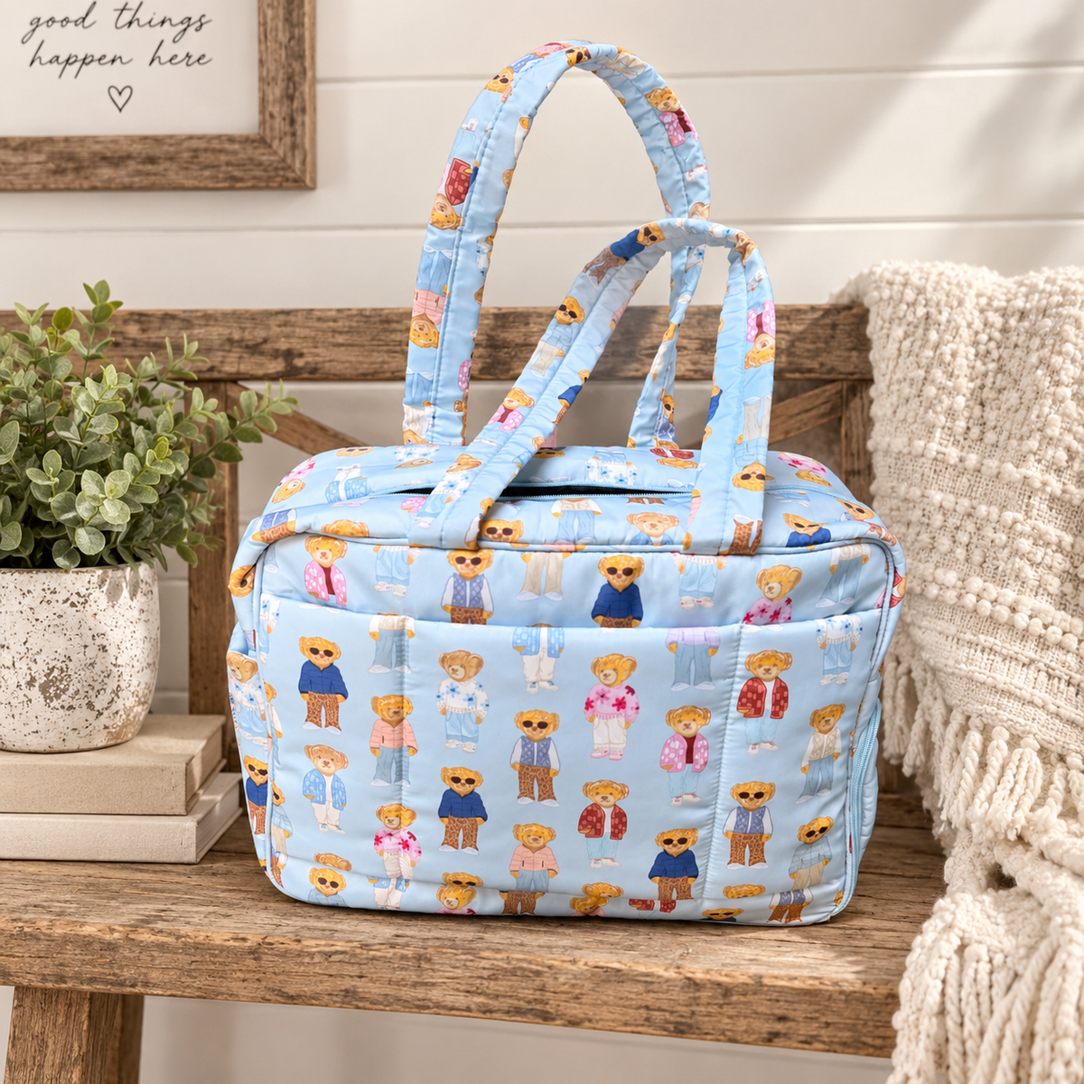 Blue Teddy Bear Weekender Bag on rustic wood bench showing light blue quilted nylon with allover teddy bear print and dual padded print handles beside speckled white pot of greenery, stacked books, and cream textured throw with Good Things Happen Here framed sign on white shiplap background
