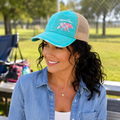 Woman wearing aqua Mama Bear distressed trucker hat with plaid bear embroidery at outdoor ball field
