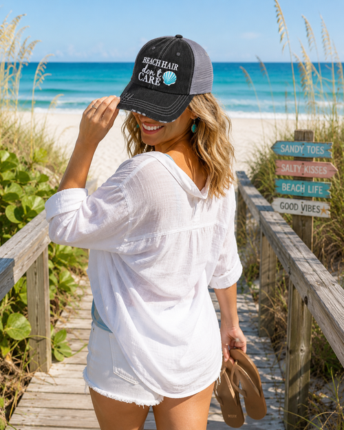A woman on a wooden boardwalk at the beach wearing a black and gray trucker hat with the text 'BEACH HAIR Don't Care' and a white tunic top, holding brown sandals.