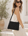 Brunette woman in white sleeveless linen top and wide-leg linen trousers wearing Black Woven Bohemian Tassel Crochet Shoulder Bag on one shoulder showing macrame cord diamond lattice pattern with black suede tassel and braided cord strap against Mediterranean whitewash exterior with olive tree and terracotta urn
