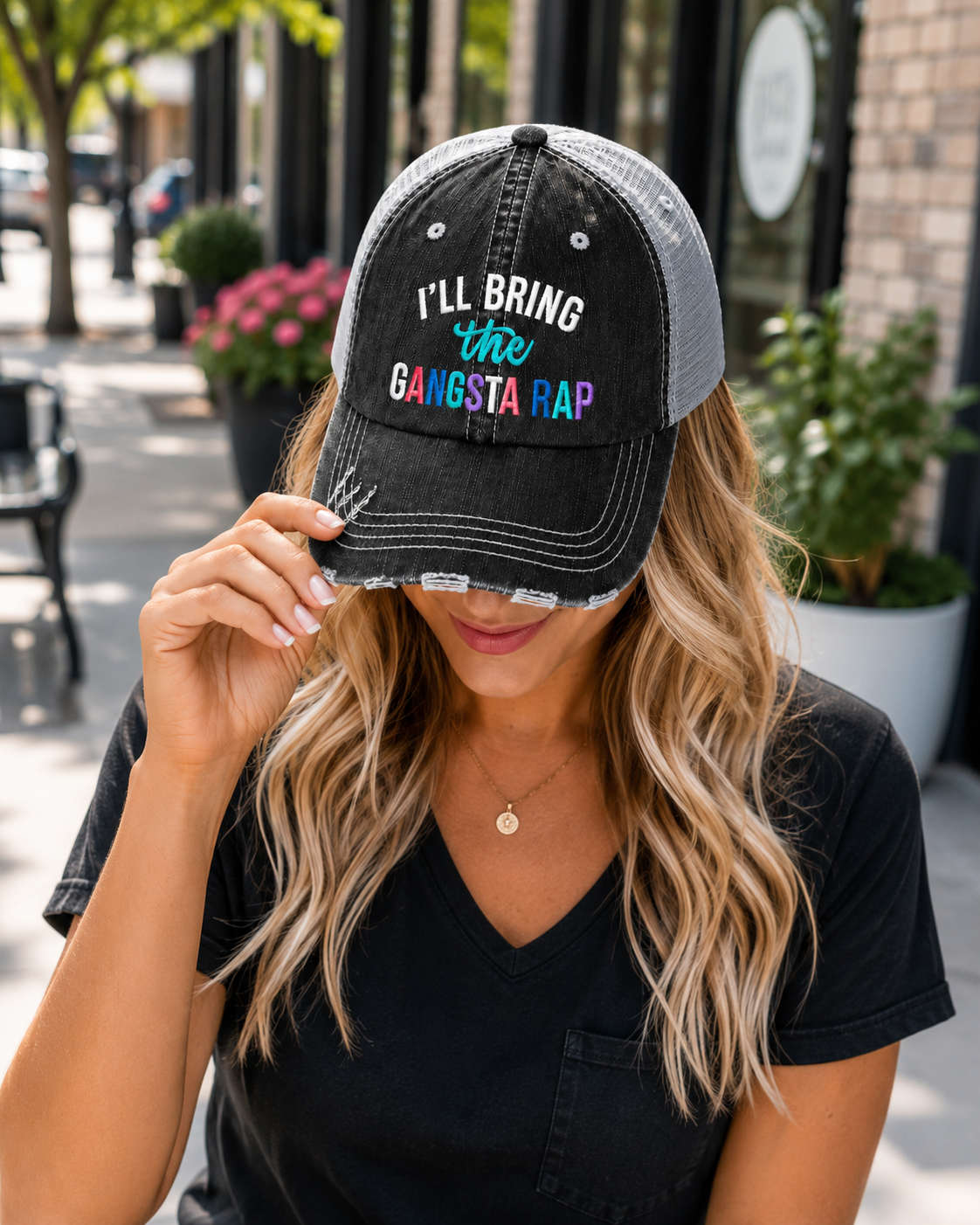 A woman wearing a black and gray trucker-style baseball cap with the text 'I'LL BRING the GANGSTA RAP' in white, teal, and pink lettering. The photo is taken outdoors on a city street with blurred storefronts and plants in the background.