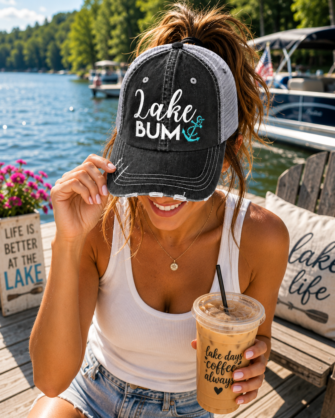 A woman sitting on a wooden dock by a lake wears a black and gray trucker-style baseball cap with 'Lake Bum' and an anchor graphic printed on the front. She is holding an iced coffee and smiling.