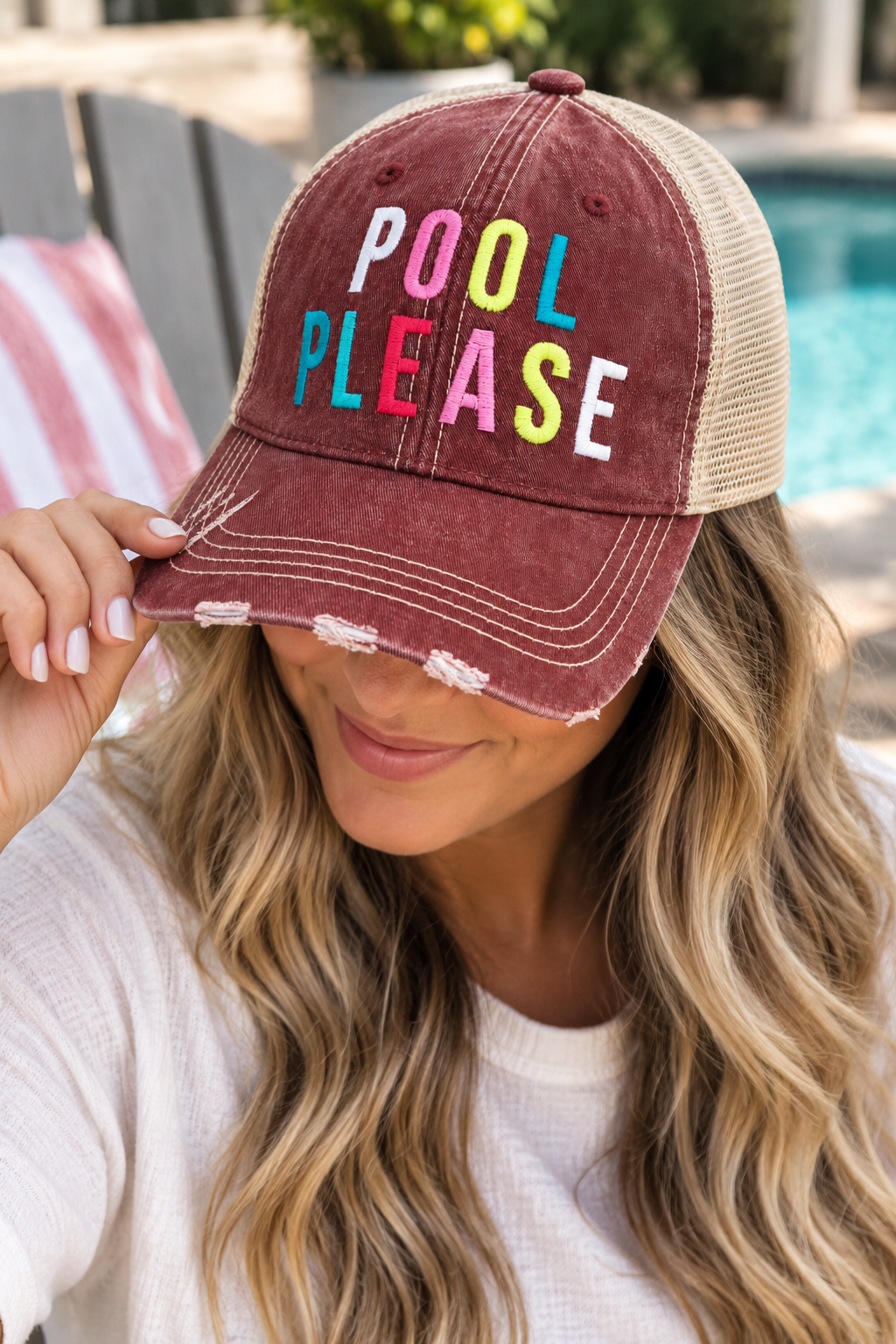 Woman wearing Pool Please Trucker Hat with distressed burgundy front and tan mesh back tilted down showing multicolor embroidered Pool Please lettering with white tee and poolside backdrop
