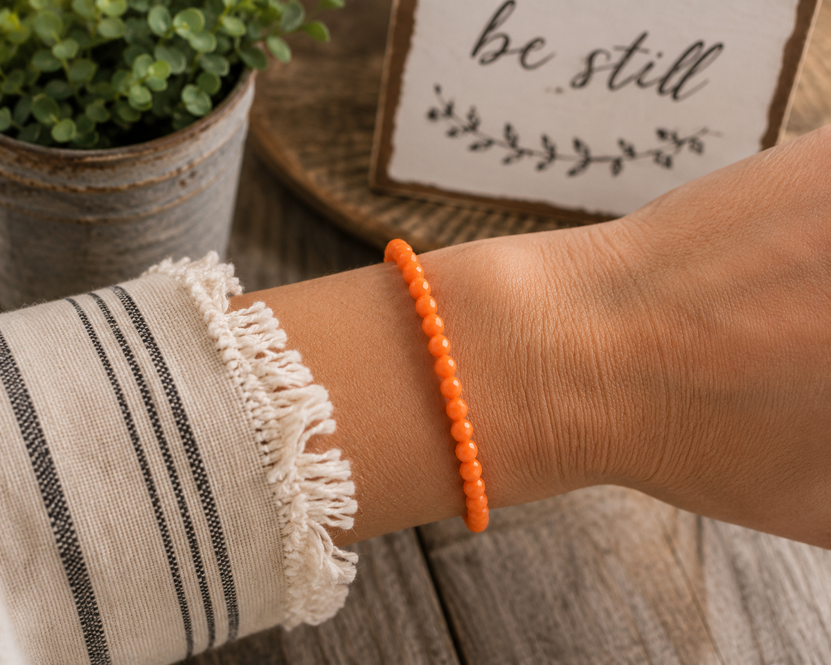 Woman wearing orange faceted bead stretch bracelet alone on wrist with white fringe top and rustic be still sign backdrop
