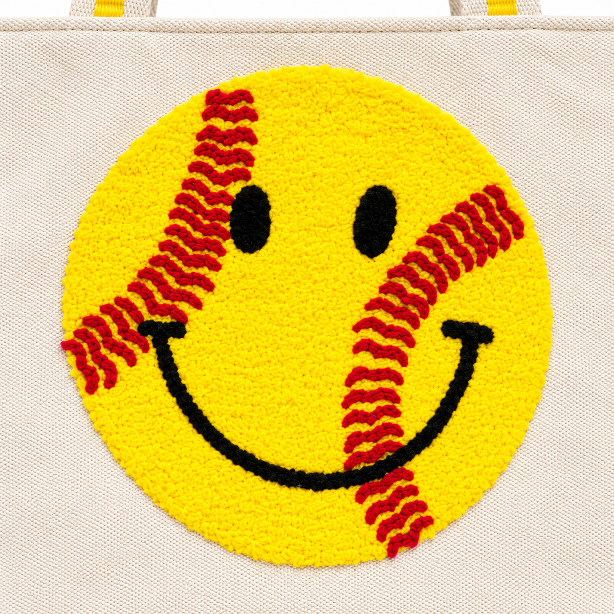 Close-up of large yellow textured punch needle softball smiley face with red curved stitching lines and black embroidered eyes and smile on natural canvas tote bag

