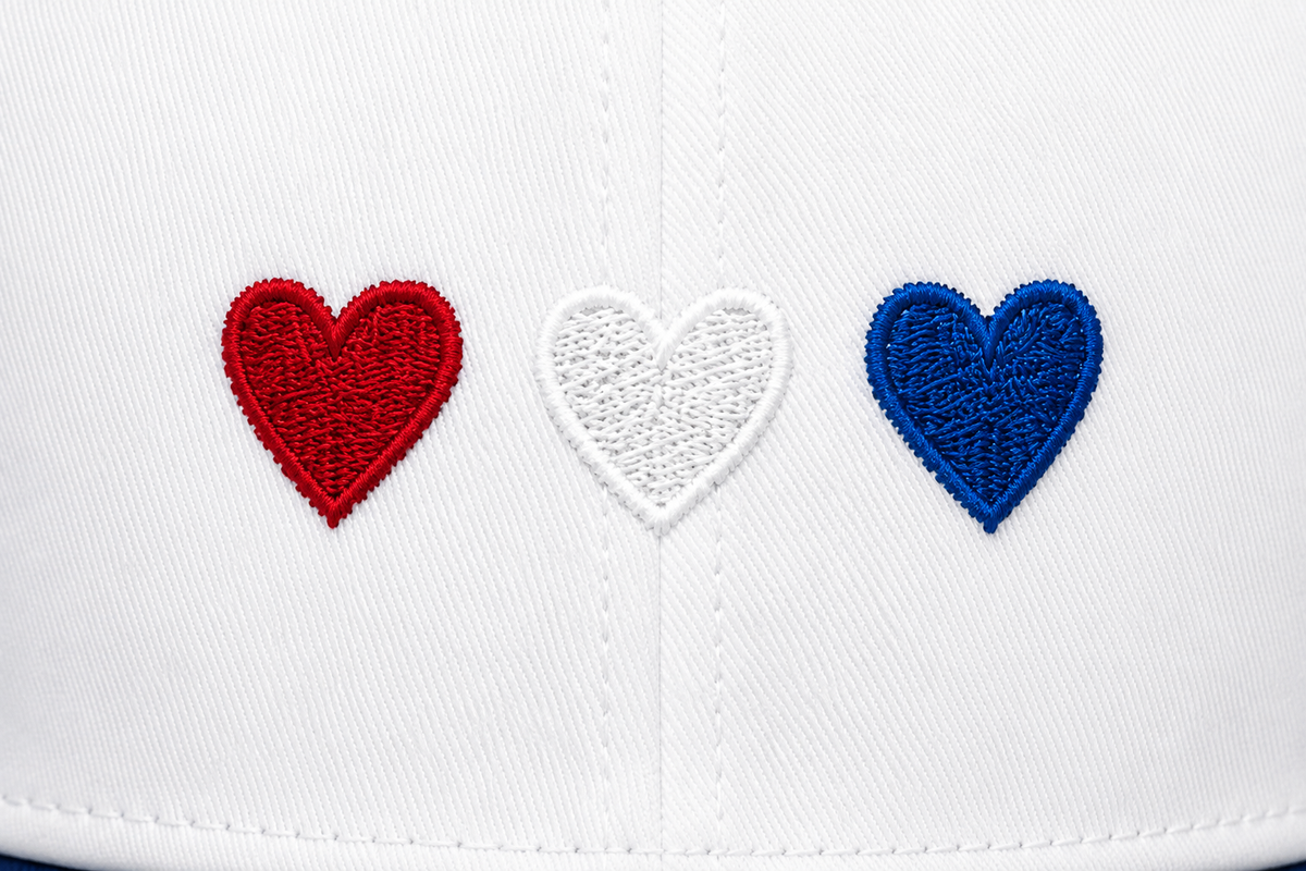 Close-up of three embroidered hearts in red, white, and blue on a white baseball cap front panel, showing detailed patriotic embroidery.

