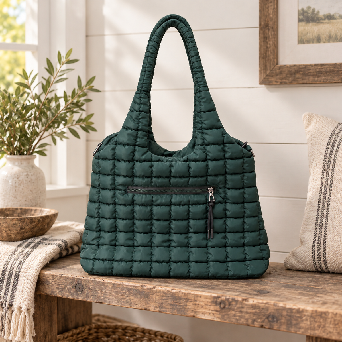 Forest Green Quilted Tote Bag on rustic wood bench showing forest green grid-quilted puffer nylon, single looped quilted shoulder strap, and front exterior zippered pocket against white shiplap wall beside white ceramic vase of olive branches, wooden bowl, striped throw blanket, and framed farmhouse landscape print

