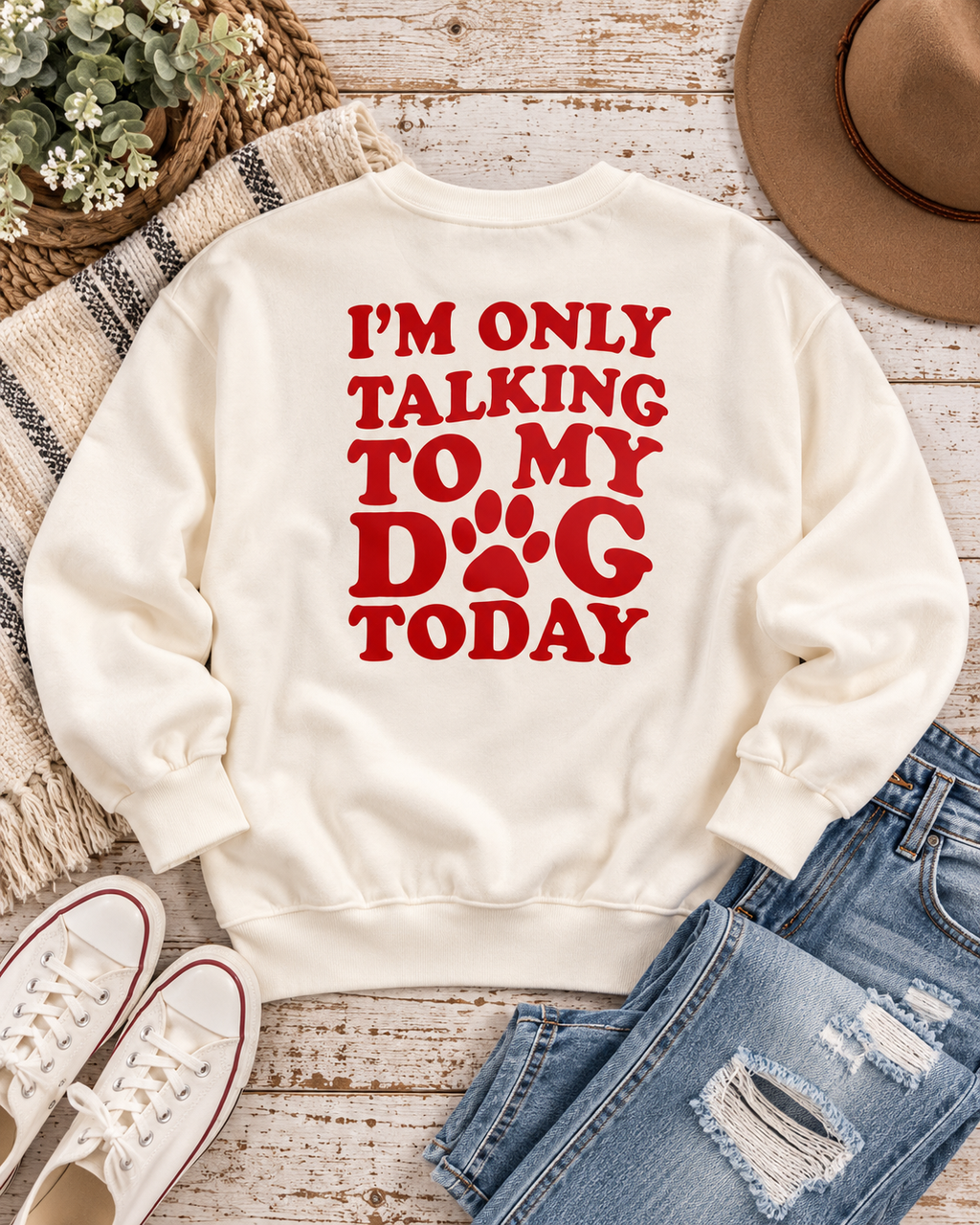 Back of the cream dog-lover sweatshirt styled with distressed jeans, white sneakers, a tan hat, and greenery on rustic wood
