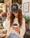 A woman sitting at an outdoor cafe wearing a dark gray and beige trucker hat with a colorful rainbow graphic on the front. She is holding a paper coffee cup and wearing a cream knit sweater.
