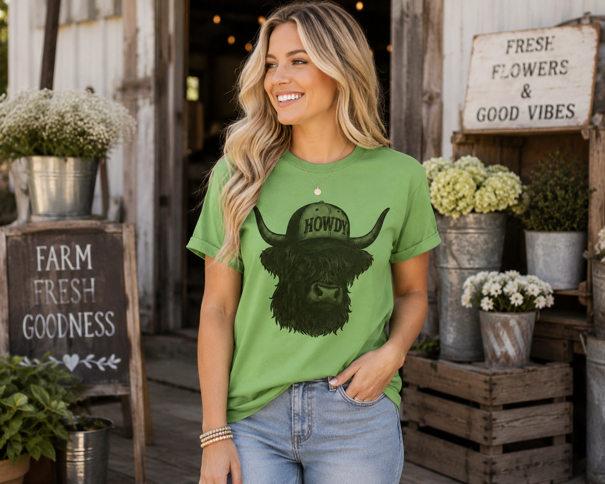A woman wearing a green graphic t-shirt with a black bull design and the word 'HOWDY' on a hat, standing in front of a rustic barn door with decorative signs and potted plants.