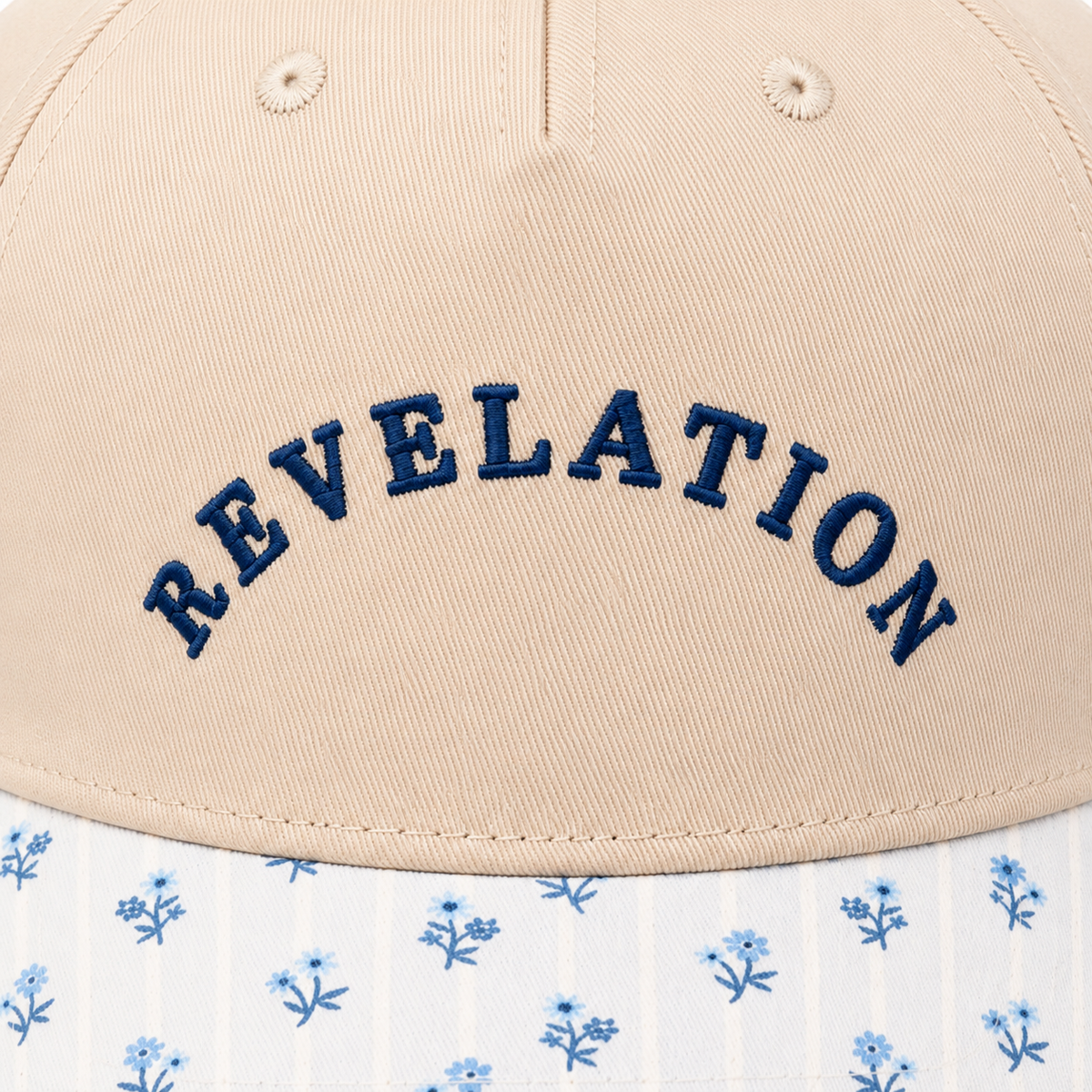 Close-up of bold navy embroidered "REVELATION" lettering in arched style on a sand baseball cap front panel with blue floral brim.

