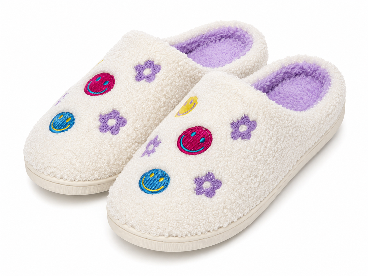 Pair of Ivory Smiley Face Fuzzy Slippers at an angle showing multicolor embroidered smiley faces and purple daisy flowers on ivory fuzzy exterior with lavender plush interior lining

