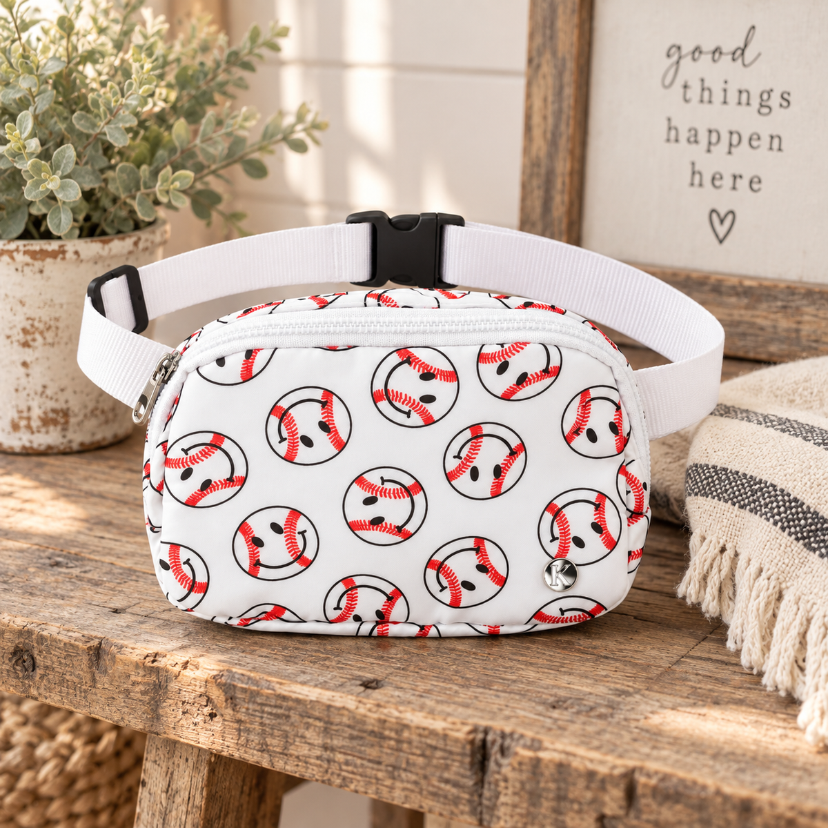Baseball Smiley Belt Bag on rustic wood bench showing white nylon with allover red and black baseball smiley face print, silver Katydid K logo rivet, and adjustable white webbing strap with black buckle beside distressed white pot of eucalyptus and striped linen throw with Good Things Happen Here sign on white shiplap background
