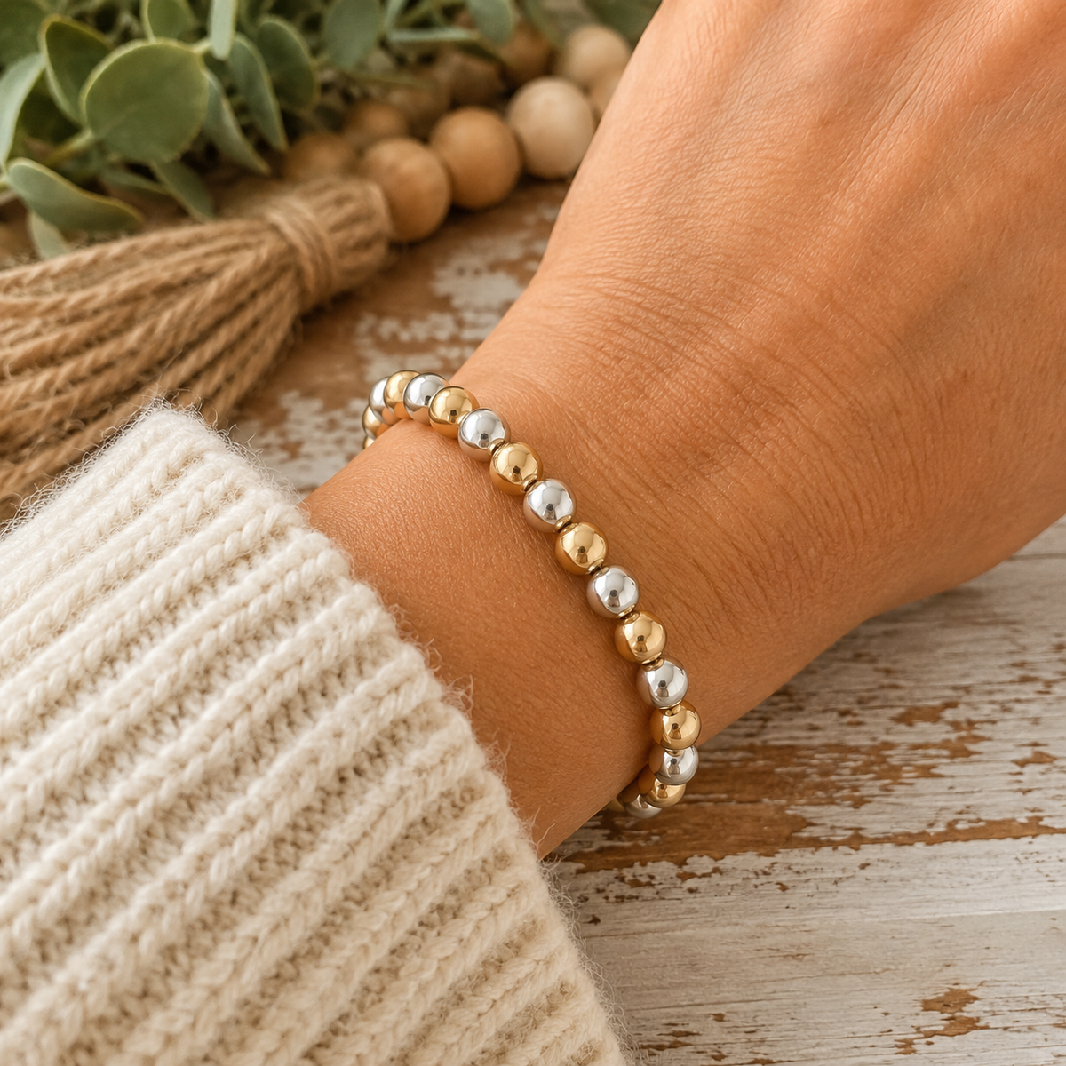 Woman wearing gold and silver round bead stretch bracelet on wrist with cream knit sweater and wood bead garland backdrop
