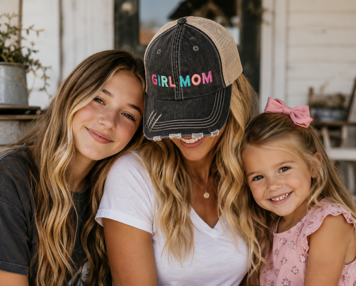 Three women posing closely together outdoors, with the central woman wearing a dark washed trucker-style baseball cap with 'GIRL MOM' embroidered on the front in pink, blue, and green letters. The woman on the left has long wavy brown hair, and the woman on the right has a pink bow in her hair and is wearing a pink floral top.